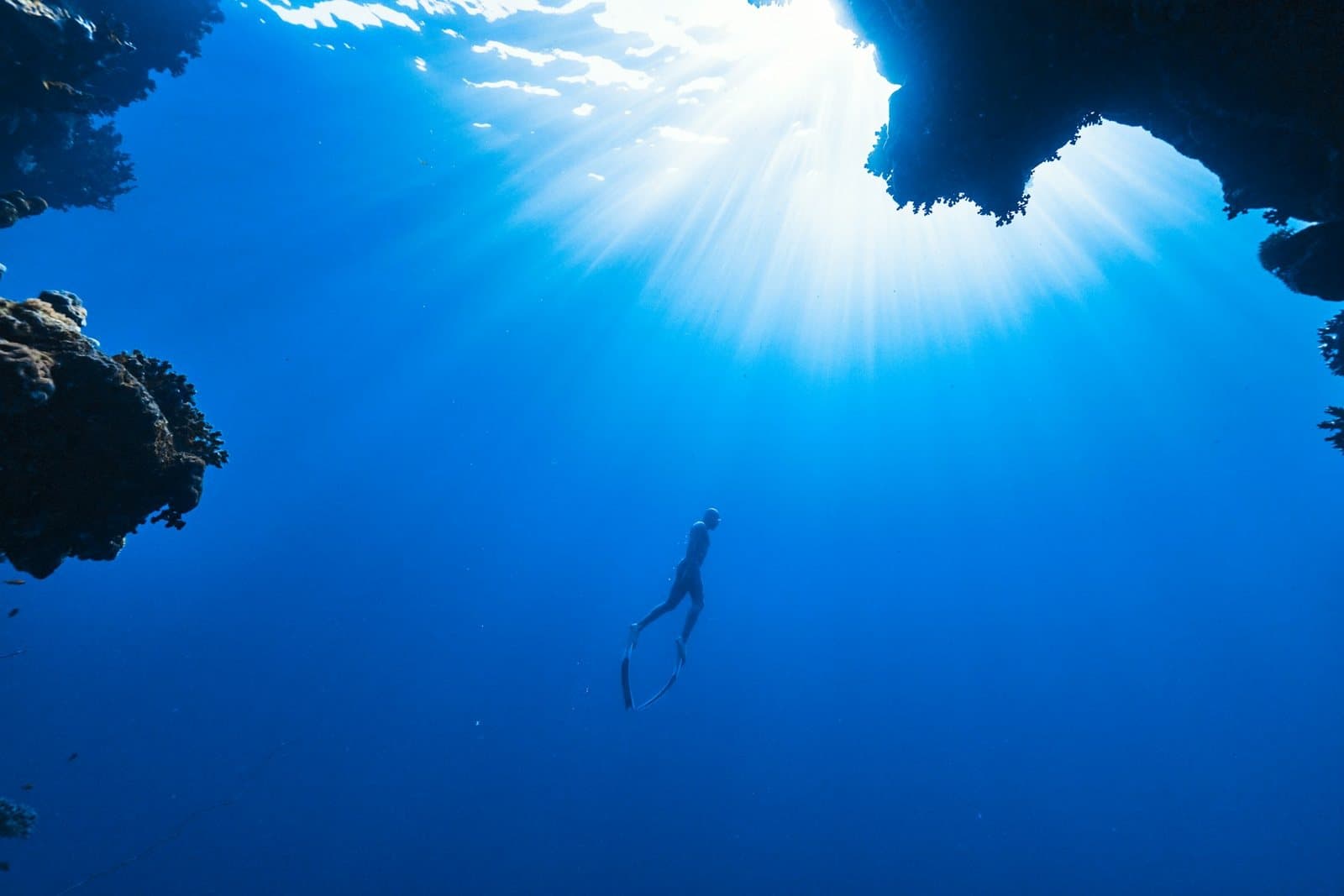 A freediver descending through clear blue water below a sunlit reef opening