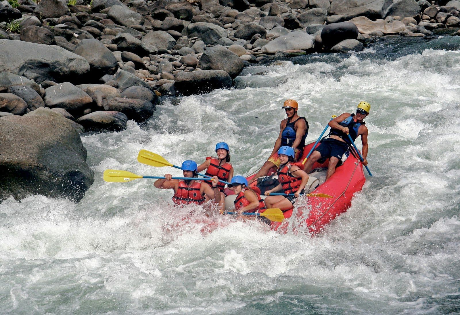 A raft crew mid river, paddles up, taking a small rapid between green banks