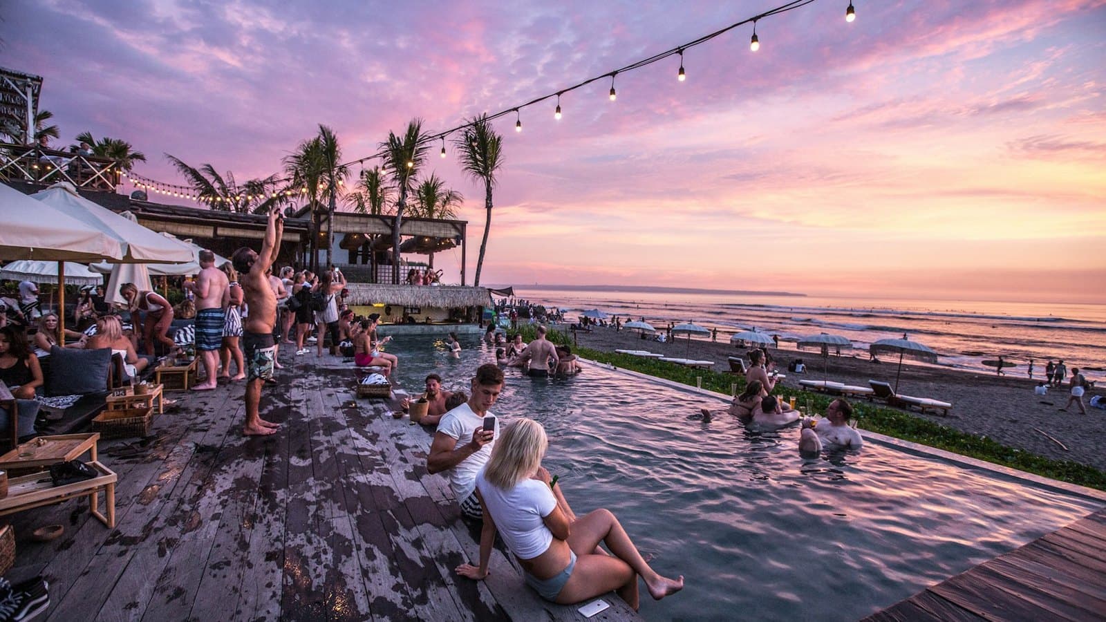 An infinity pool crowded with people at sunset, the sea beyond it and festoon lights overhead