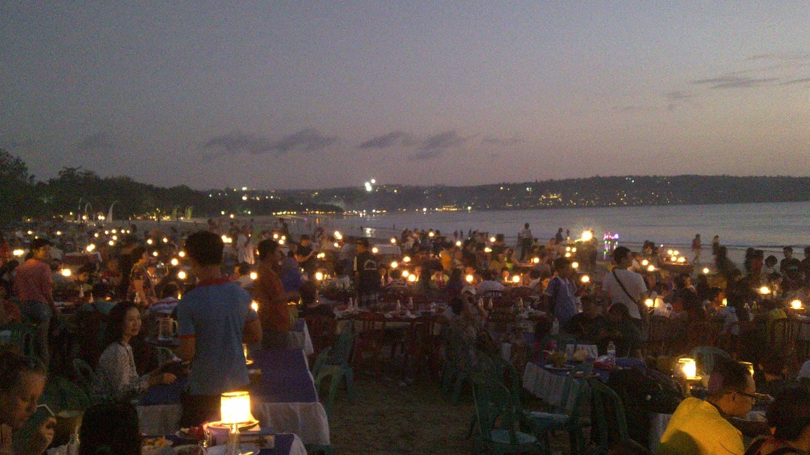 Rows of candlelit tables set out on Jimbaran beach at dusk, the bay and the lights of the airport road behind