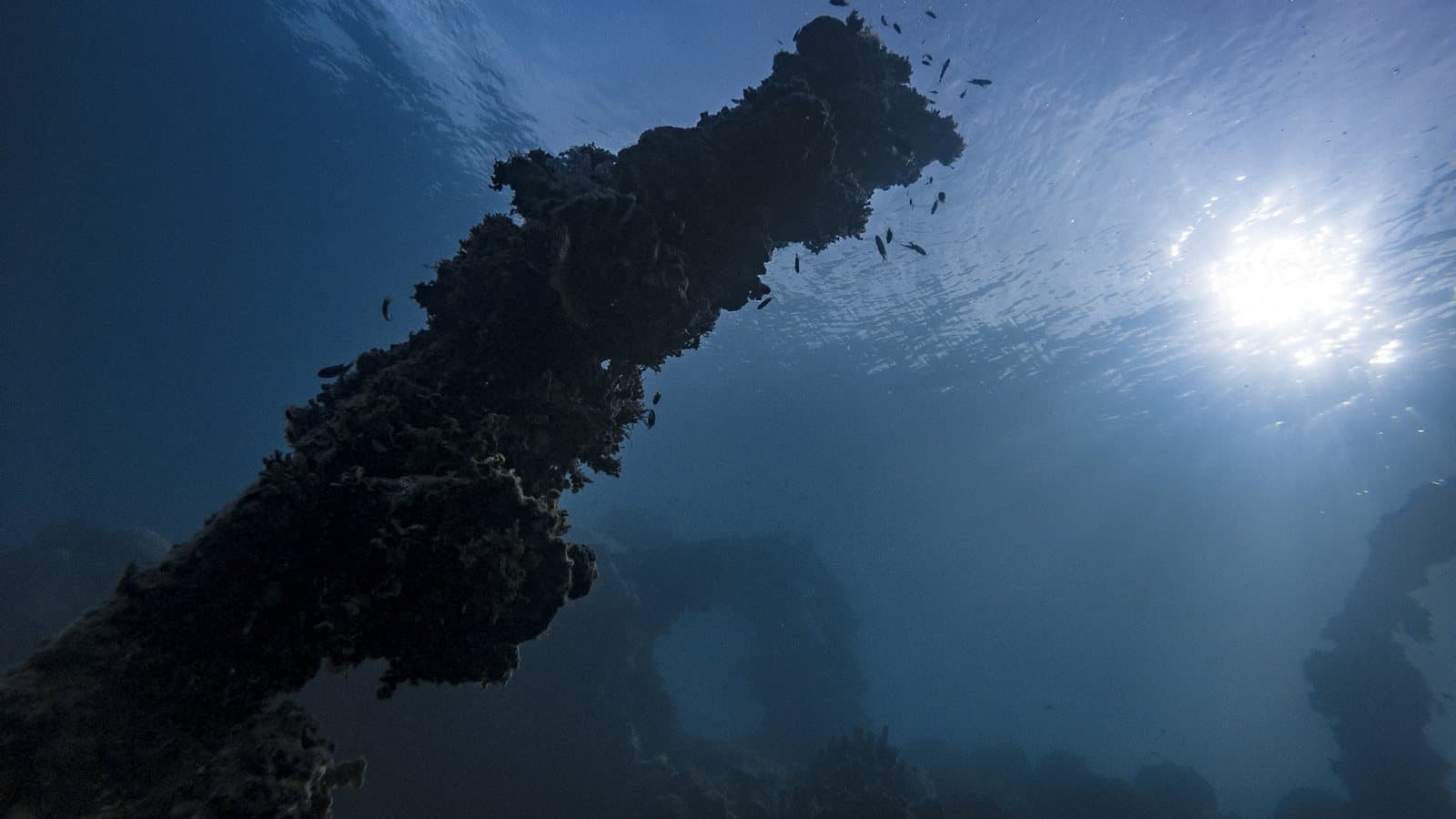 The corroded hull of a sunken ship rising through clear water toward the surface light