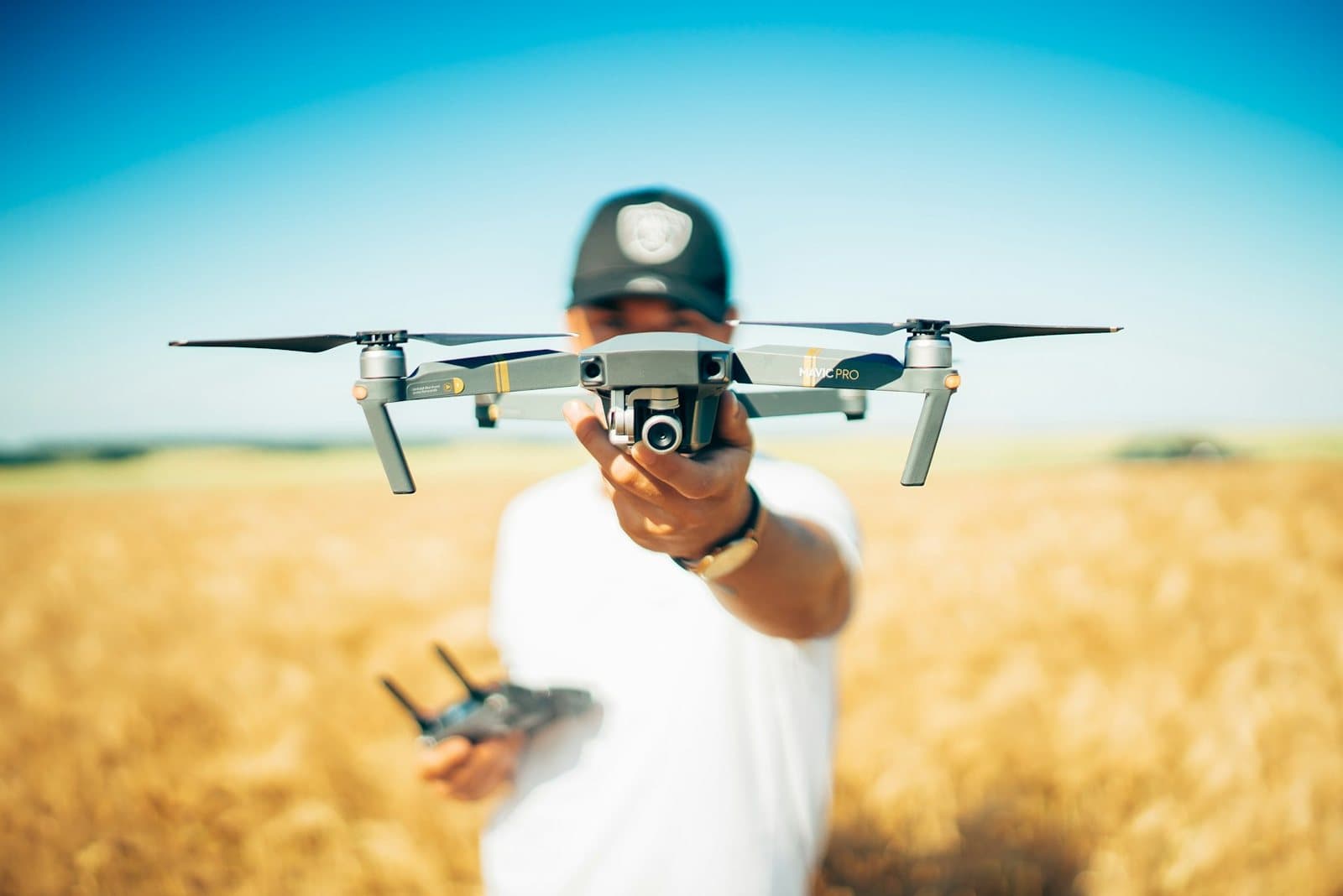 A drone pilot holding a folded quadcopter at eye level in an open field, controller in the other hand
