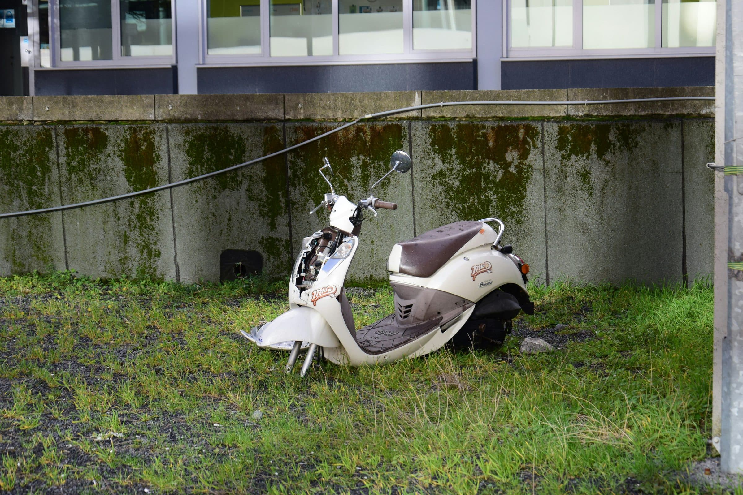 A white scooter left on grass against a concrete wall, its front fairing broken and its panels cracked