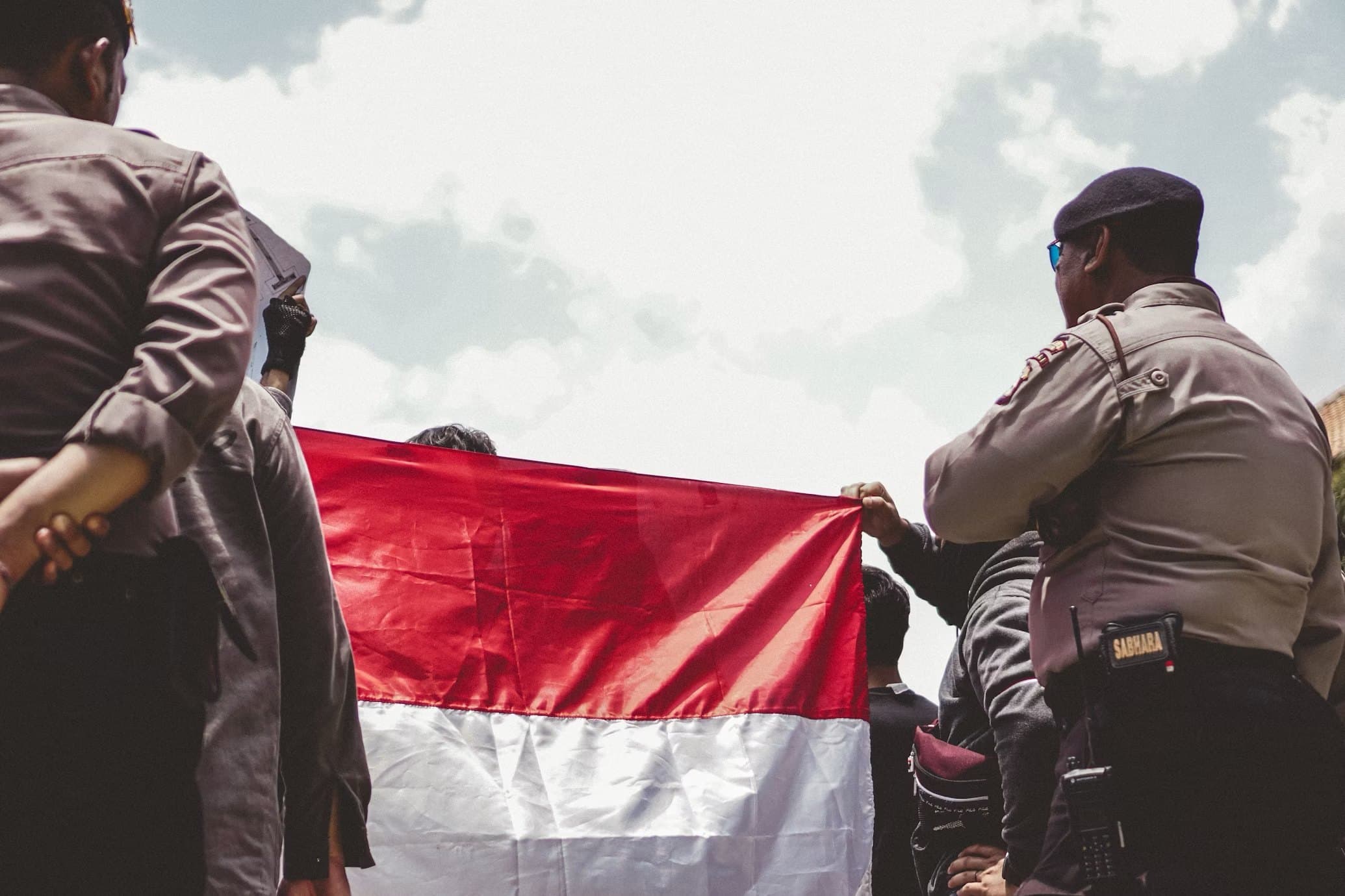 Indonesian police officers in service uniform standing either side of a national flag held up in a crowd