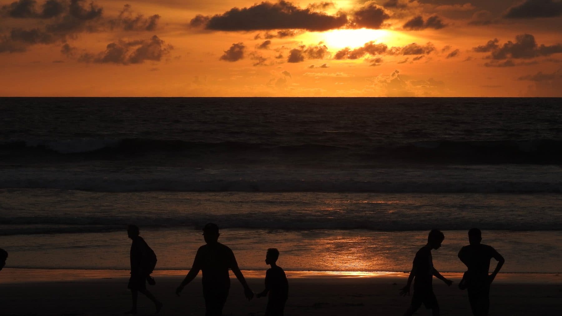 People standing at the water line on Legian beach, silhouetted against a low sun and a band of cloud