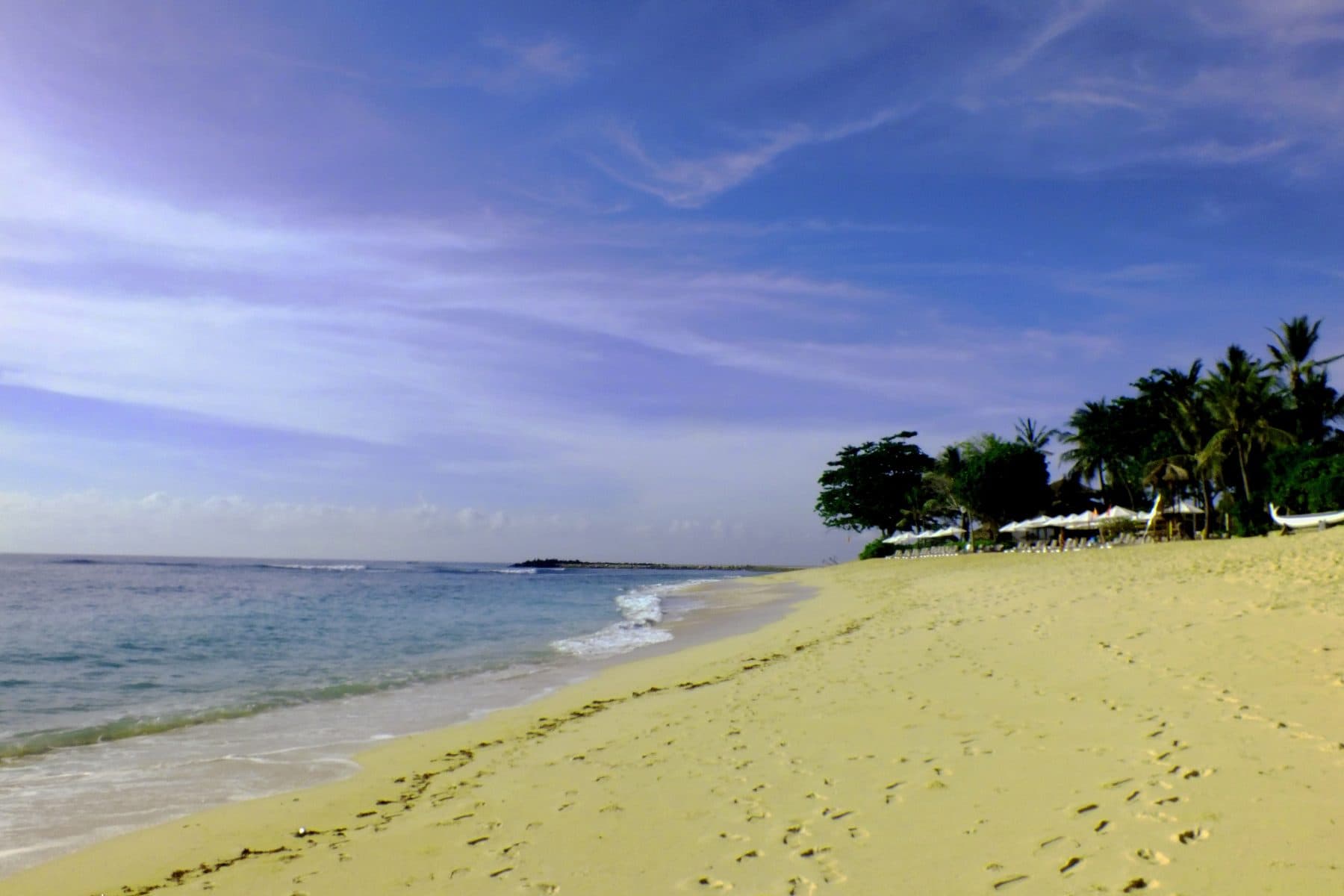 The pale sand of Nusa Dua beach curving towards a headland of palms, with calm shallow water