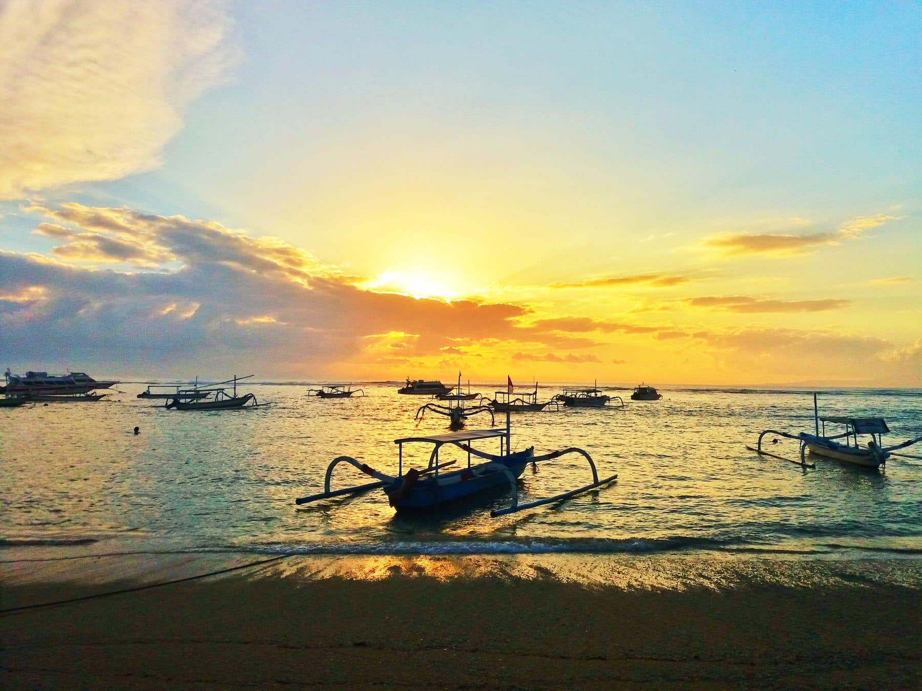A traditional jukung outrigger moored off Sanur beach at sunrise, its hull dark against the low sun