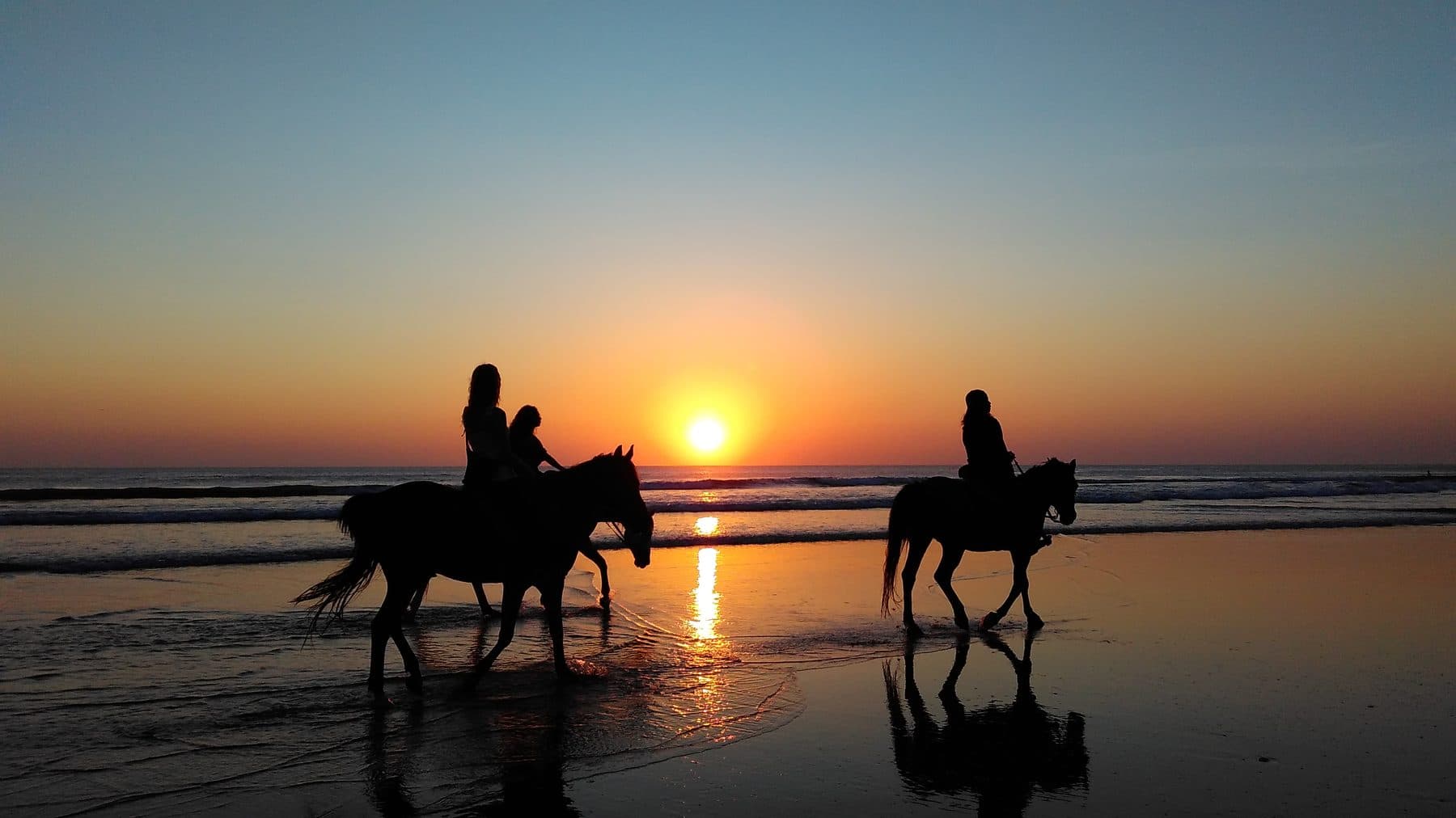 Three riders on horseback silhouetted against the sun on the wet sand of a Seminyak beach at dusk